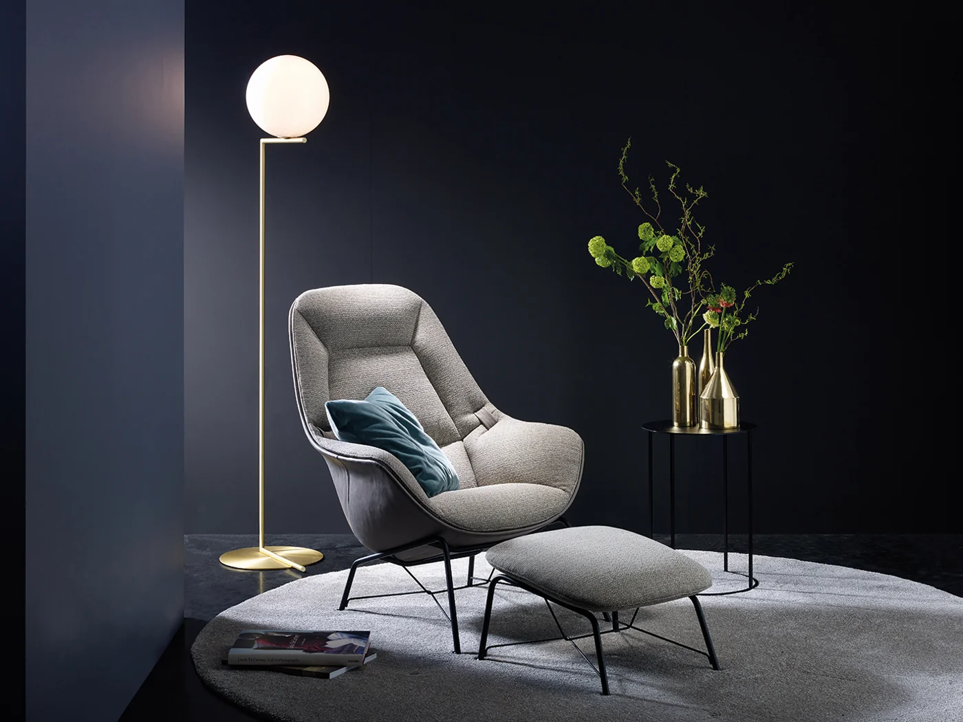 Grauer JORI Prelude Lounge Sessel mit dekorativen Bändern und Hocker vor dunkler Wand