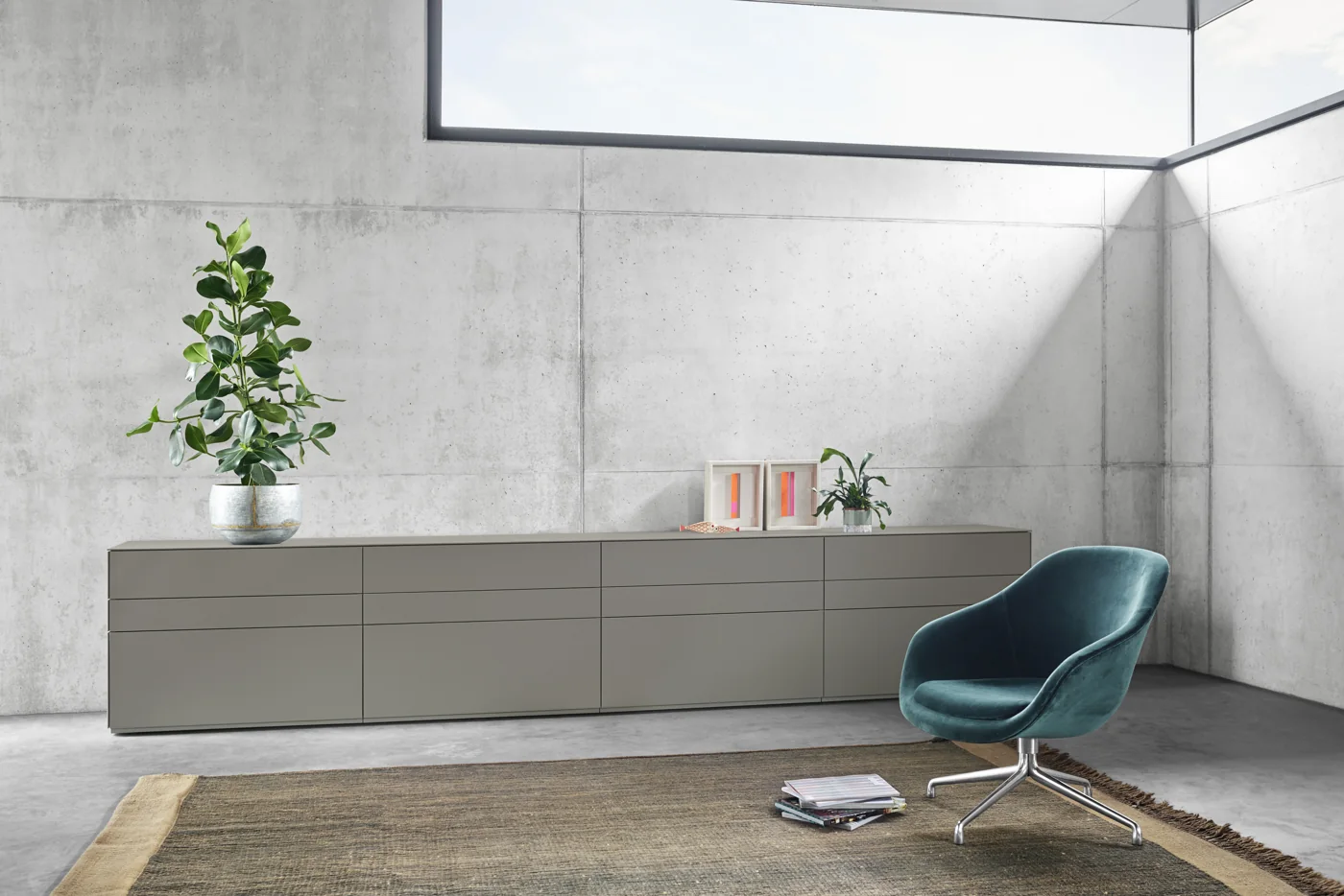 Langes graugrünes Kettnaker SOMA-Sideboard vor einer Betonwand neben einem blauen Sessel