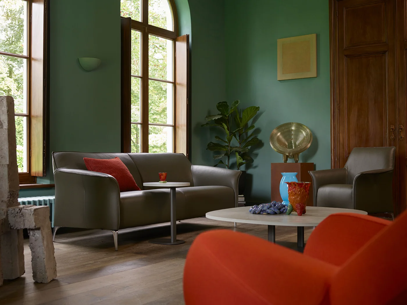 Mayon Sofa in dunklem Bezug mit rotem Mayuro Sessel im Vordergrund